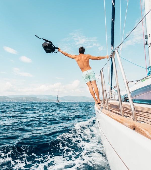 A photo of a man in swim trunks standing on the edge of a boat out at sea, arm outstretched, holding a backpack.