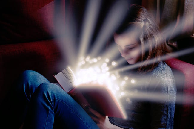 A photo of a young girl reading a book that has a magical light emanating from its pages.